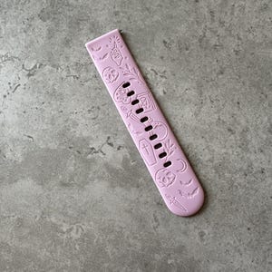 May include: A pink silicone watch band with a Halloween theme. The band is decorated with embossed images of bats, skulls, coffins, and other spooky symbols.