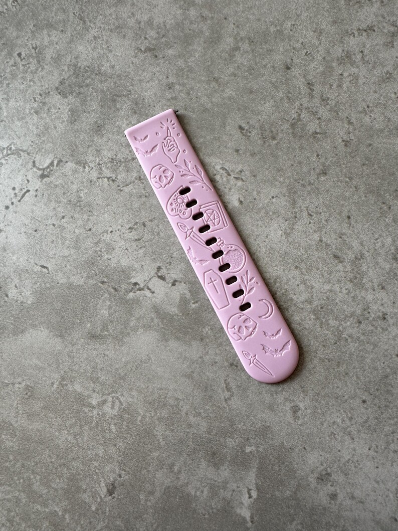May include: A pink silicone watch band with a Halloween theme. The band is decorated with embossed images of bats, skulls, coffins, and other spooky symbols.