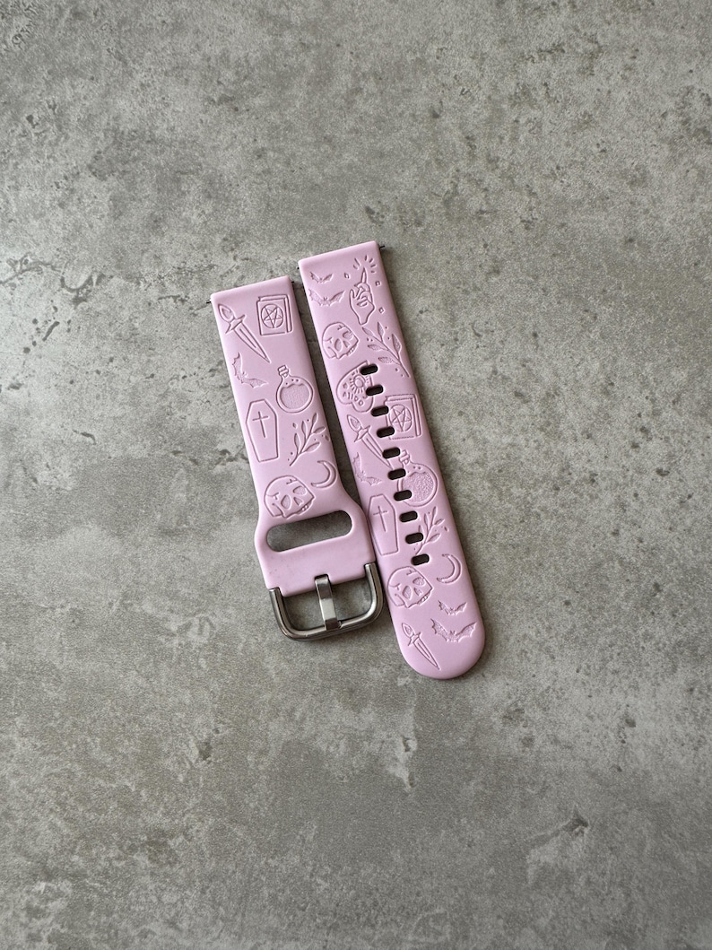 May include: A pair of pink silicone watch bands with a Halloween theme. The bands feature embossed designs of skulls, bats, crosses, daggers, and other spooky symbols.