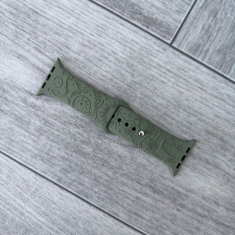 K&ouml;nnte beinhalten: Ein olivgr&uuml;nes Silikon-Uhrenarmband mit einem eingravierten Muster aus himmlischen Elementen wie Sonne, Mond, Sternen und floralen Motiven. Das Armband verf&uuml;gt &uuml;ber einen Metallstiftverschluss und mehrere Einstelll&ouml;cher f&uuml;r eine sichere Passform.