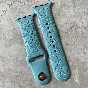 K&ouml;nnte beinhalten: Ein mattes, staubblaues Silikon-Uhrenarmband mit gepr&auml;gten Motiven aus Himmelsk&ouml;rpern und Natur. Das Muster zeigt Sonne, Mond, Sterne, eine Schlange, Blumen und Motten. Das Armband verf&uuml;gt &uuml;ber einen Metallstiftverschluss und Standardanschl&uuml;sse.