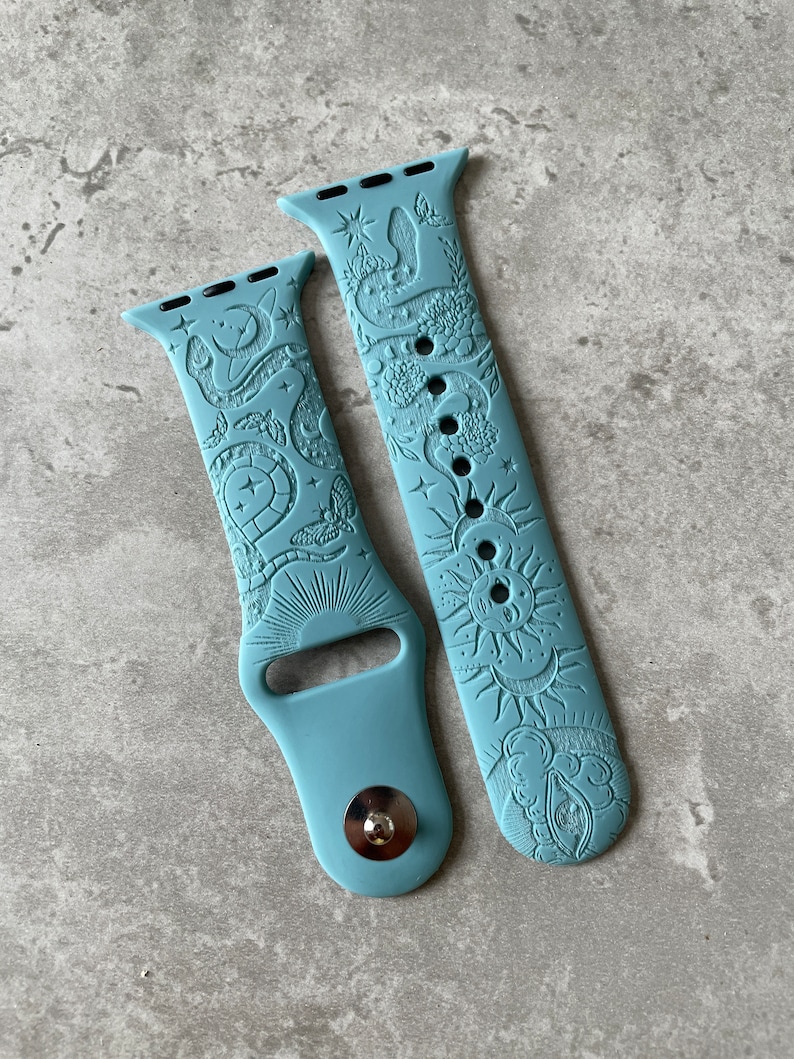 K&ouml;nnte beinhalten: Ein mattes, staubblaues Silikon-Uhrenarmband mit gepr&auml;gten Motiven aus Himmelsk&ouml;rpern und Natur. Das Muster zeigt Sonne, Mond, Sterne, eine Schlange, Blumen und Motten. Das Armband verf&uuml;gt &uuml;ber einen Metallstiftverschluss und Standardanschl&uuml;sse.