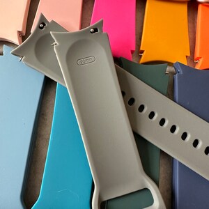 May include: A collection of colorful 20mm watch bands in various shades, including pink, orange, blue, and gray. The bands are made of a flexible material and feature a buckle closure. The gray band has "20mm" printed on it.