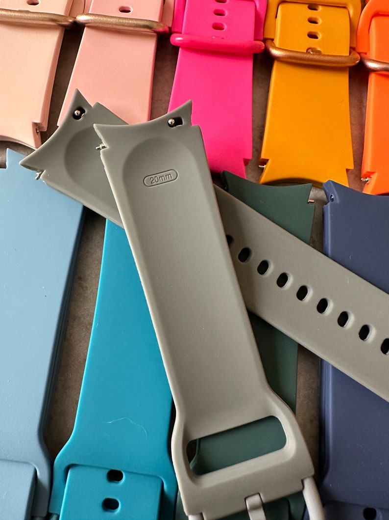 May include: A collection of colorful 20mm watch bands in various shades, including pink, orange, blue, and gray. The bands are made of a flexible material and feature a buckle closure. The gray band has "20mm" printed on it.