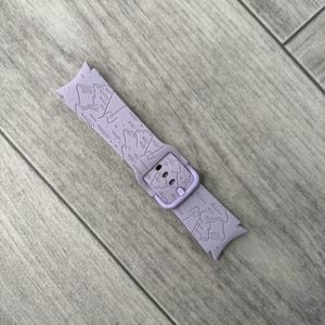 May include: A lavender watch band with a mountain and tree design. The band features a buckle and is made of a flexible material. The mountain design is etched into the band, creating a textured look. The band is shown on a wooden surface.