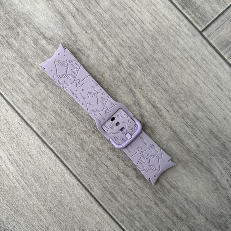 May include: A lavender watch band with a mountain and tree design. The band features a buckle and is made of a flexible material. The mountain design is etched into the band, creating a textured look. The band is shown on a wooden surface.