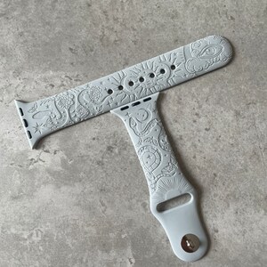 K&ouml;nnte beinhalten: Ein hellgraues Silikon-Uhrenarmband mit gepr&auml;gtem Himmelsmotiv. Das Muster zeigt eine Sonne mit Gesicht, Mondsicheln, Sterne, Schlangen, Motten und Blumen. Das Armband verf&uuml;gt &uuml;ber einen Metallstiftverschluss und Standard-Anschlussst&uuml;cke.