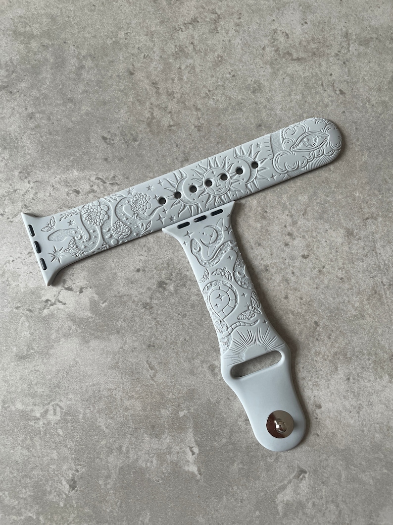 K&ouml;nnte beinhalten: Ein hellgraues Silikon-Uhrenarmband mit gepr&auml;gtem Himmelsmotiv. Das Muster zeigt eine Sonne mit Gesicht, Mondsicheln, Sterne, Schlangen, Motten und Blumen. Das Armband verf&uuml;gt &uuml;ber einen Metallstiftverschluss und Standard-Anschlussst&uuml;cke.
