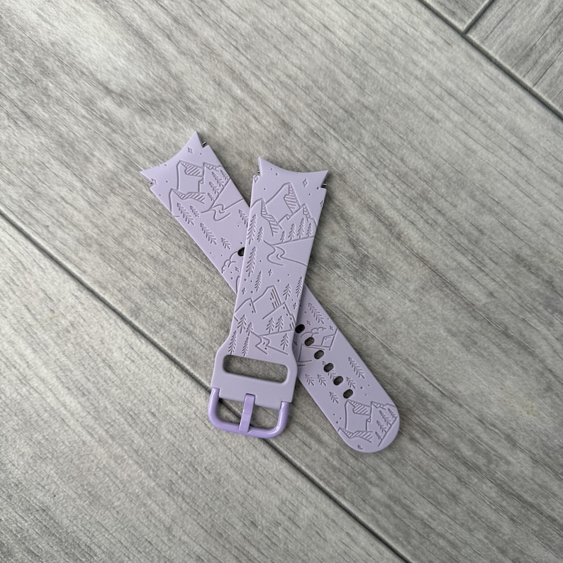 May include: Two lavender watch bands with a mountain and river design. The bands are made of a soft material and feature a buckle closure. The bands are designed to fit a smartwatch and are a light purple color.
