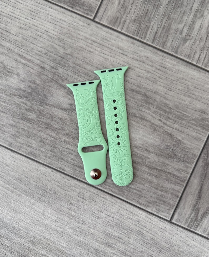 K&ouml;nnte beinhalten: Ein helles mintgr&uuml;nes Silikon-Uhrenarmband mit einem gepr&auml;gten Muster aus Schlangen, Sonnen, Monden, Sternen und einem Auge. Das zweiteilige Armband verf&uuml;gt &uuml;ber einen Metallstiftverschluss und mehrere L&ouml;cher zur individuellen Anpassung.