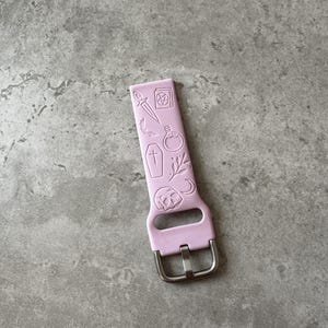May include: A light pink silicone watch band with a silver buckle. The band has a design of various occult symbols, including a pentagram, a dagger, a coffin, a potion bottle, and a crescent moon.