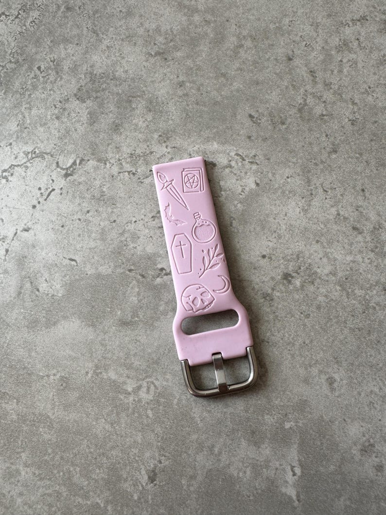 May include: A light pink silicone watch band with a silver buckle. The band has a design of various occult symbols, including a pentagram, a dagger, a coffin, a potion bottle, and a crescent moon.