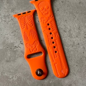 Könnte beinhalten: Zwei orangefarbene Silikon-Apple Watch-Armbänder mit Skelett-Design. Ein Armband zeigt einen Schädel und Knochen, während das andere ein vollständiges Skelett zeigt.