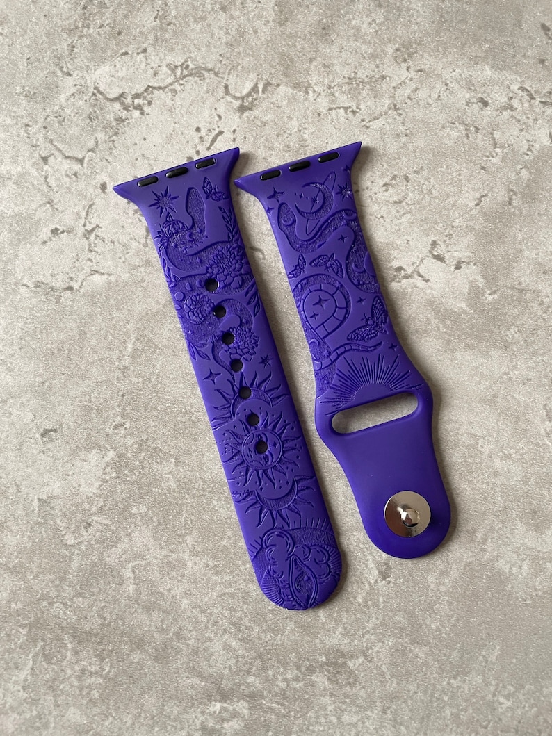 K&ouml;nnte beinhalten: Ein violettes Silikon-Uhrenarmband mit gepr&auml;gtem Muster aus Schlangen, Motten, Sonnen, Monden und Sternen. Das Armband besteht aus zwei Teilen, einem mit mehreren Einstelll&ouml;chern und einem mit einem silberfarbenen Metallstift-Verschlussmechanismus.