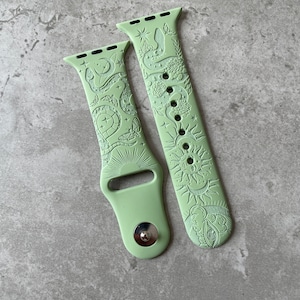 K&ouml;nnte beinhalten: Ein helles salbeigr&uuml;nes Silikon-Uhrenarmband mit gepr&auml;gtem Himmelsmotiv. Das Muster zeigt eine Sonne mit Gesicht, Mondsicheln, Sterne, Motten und eine Schlange, die sich durch florale Elemente windet. Das Armband hat einen Metallverschluss.