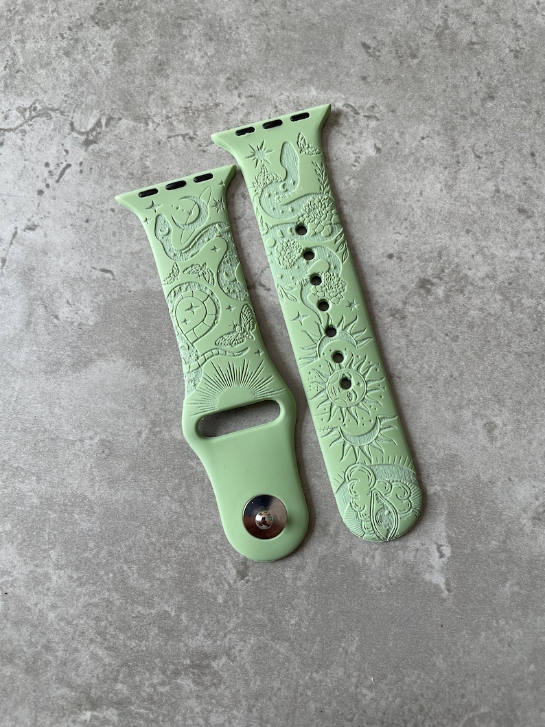 K&ouml;nnte beinhalten: Ein helles salbeigr&uuml;nes Silikon-Uhrenarmband mit gepr&auml;gtem Himmelsmotiv. Das Muster zeigt eine Sonne mit Gesicht, Mondsicheln, Sterne, Motten und eine Schlange, die sich durch florale Elemente windet. Das Armband hat einen Metallverschluss.