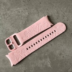 May include: A pink silicone watch strap with a floral pattern. The strap is designed for a smartwatch and has a buckle fastening.