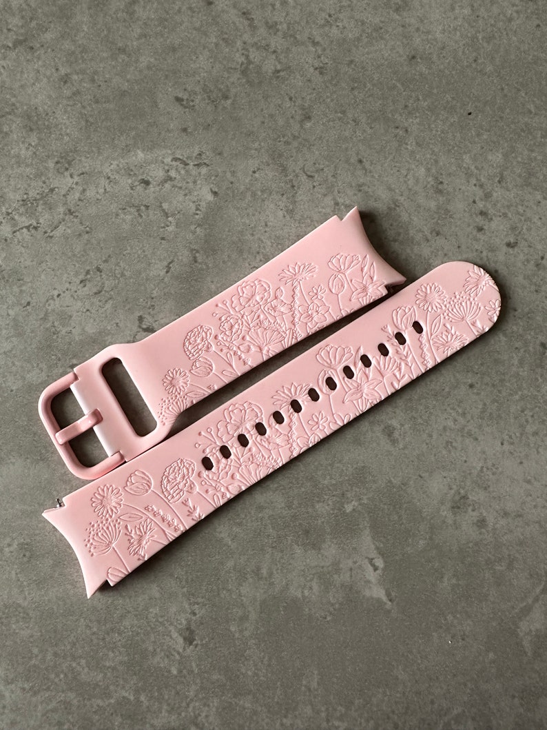 May include: A pink silicone watch strap with a floral pattern. The strap is designed for a smartwatch and has a buckle fastening.