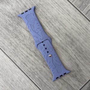 K&ouml;nnte beinhalten: Ein periwinkelfarbenes Uhrenarmband mit einem eingravierten Design von Walhaien und Fischen. Das Armband besteht aus einem flexiblen Material und hat einen silberfarbenen Verschluss. Das Design ist &uuml;ber das gesamte Armband hinweg einheitlich.