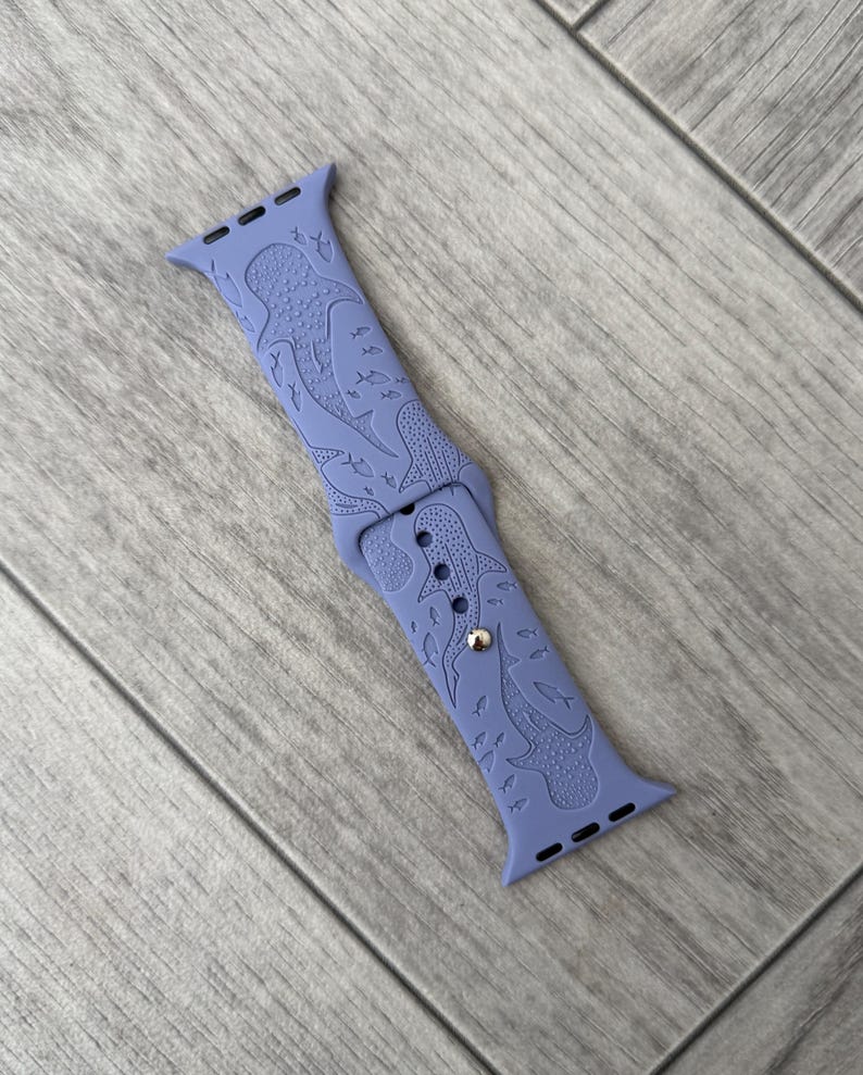K&ouml;nnte beinhalten: Ein periwinkelfarbenes Uhrenarmband mit einem eingravierten Design von Walhaien und Fischen. Das Armband besteht aus einem flexiblen Material und hat einen silberfarbenen Verschluss. Das Design ist &uuml;ber das gesamte Armband hinweg einheitlich.