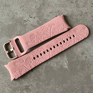 May include: A pink silicone watch strap with a floral design. The strap has a gold metal buckle.