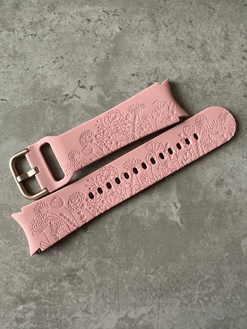 May include: A pink silicone watch strap with a floral design. The strap has a gold metal buckle.