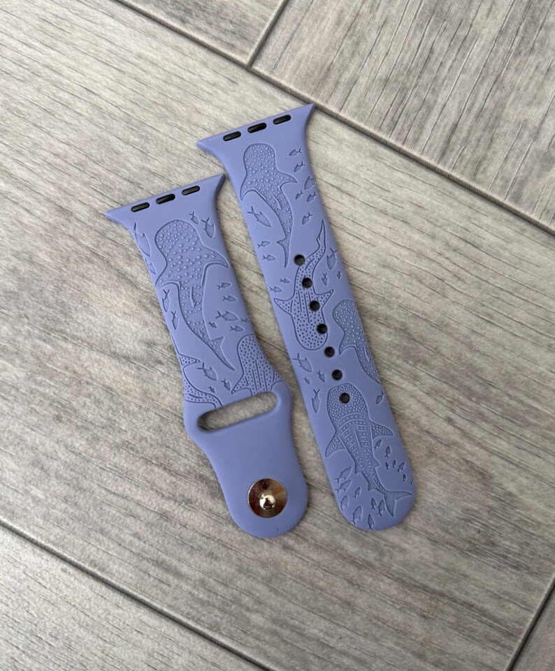 K&ouml;nnte beinhalten: Zwei lavendelfarbene Uhrenarmb&auml;nder mit einem Walhai- und Fischdesign. Die Armb&auml;nder bestehen aus einem flexiblen Material und weisen ein detailliertes gepr&auml;gtes Muster auf. Sie sind f&uuml;r eine Smartwatch konzipiert und haben einen Metallverschluss.