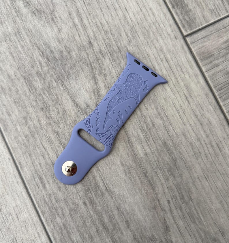 K&ouml;nnte beinhalten: Ein periwinkelfarbenes Uhrenarmband mit einem detaillierten Design von Meereskreaturen. Das Armband besteht aus einem flexiblen Material und verf&uuml;gt &uuml;ber einen silbernen Knopfverschluss. Das Design umfasst einen Oktopus und andere Meereslebewesen.