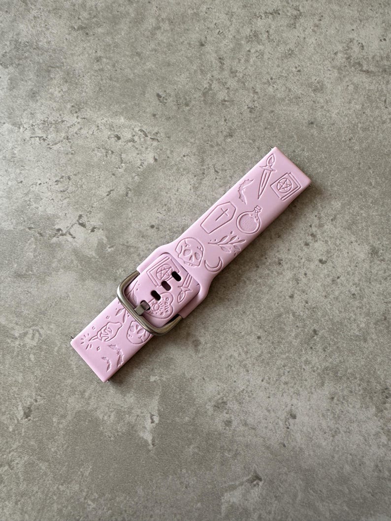 May include: A pink silicone watch band with a silver buckle. The band is decorated with a variety of small, embossed designs, including a skull, a coffin, a dagger, and a potion bottle.