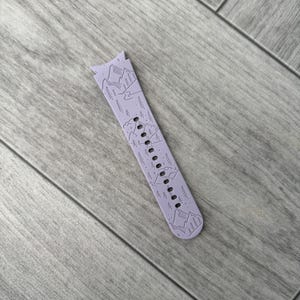 May include: A lavender watch band with a mountain and tree design. The band has a series of small holes for adjustment and is made of a flexible material. The design features a detailed illustration of mountains and trees.