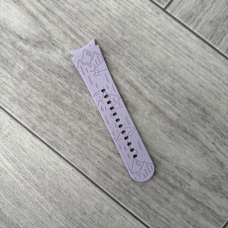 May include: A lavender watch band with a mountain and tree design. The band has a series of small holes for adjustment and is made of a flexible material. The design features a detailed illustration of mountains and trees.
