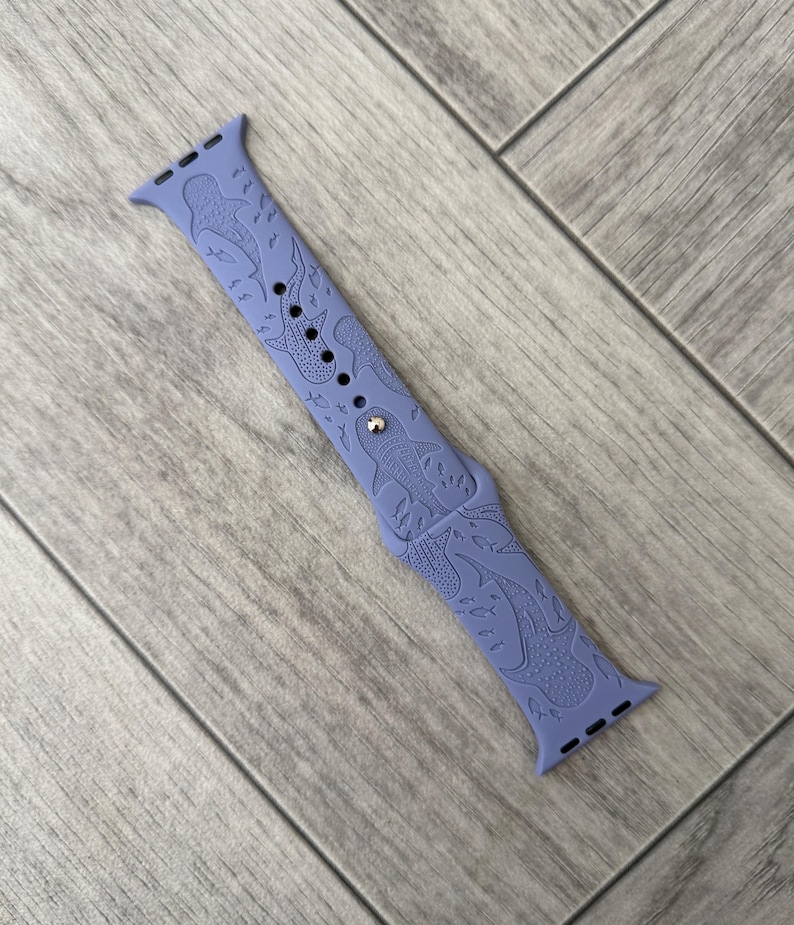 K&ouml;nnte beinhalten: Ein periwinkelfarbenes Uhrenarmband mit einem sich wiederholenden Muster aus Walhaien und anderen Meereslebewesen. Das Armband hat eine silberfarbene Schnalle und besteht aus einem flexiblen Material. Das Armband ist f&uuml;r eine Smartwatch konzipiert.