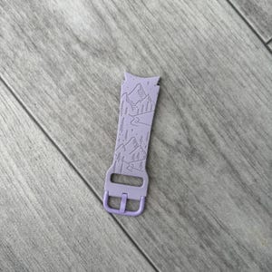 May include: A lavender watch band with a mountain and forest design. The band features a buckle and a detailed landscape illustration of mountains, trees, and a river. The band is made of a flexible material.