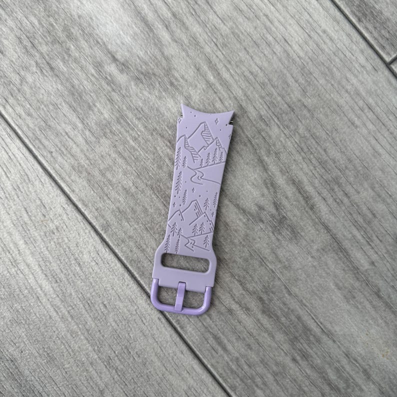 May include: A lavender watch band with a mountain and forest design. The band features a buckle and a detailed landscape illustration of mountains, trees, and a river. The band is made of a flexible material.