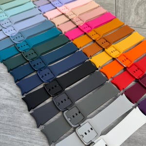 May include: A collection of colorful watch bands in various shades, including blue, pink, orange, and gray. Each band features a buckle closure and is arranged in a diagonal pattern. The bands are made of a flexible material.