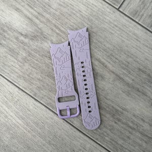 May include: A pair of lavender watch bands with a mountain and forest design. The bands are made of a flexible material and feature a buckle closure. The design includes mountains, trees, and a river, creating a nature-inspired aesthetic.