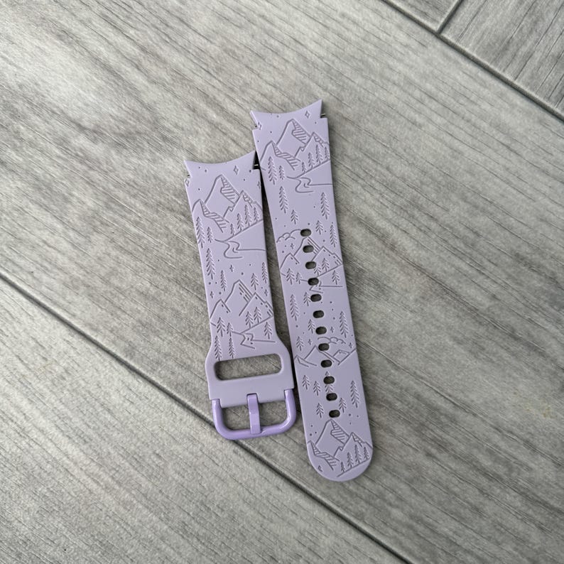 May include: A pair of lavender watch bands with a mountain and forest design. The bands are made of a flexible material and feature a buckle closure. The design includes mountains, trees, and a river, creating a nature-inspired aesthetic.