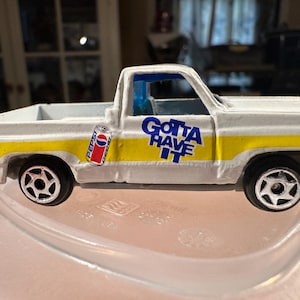 May include: A white toy pickup truck with yellow and blue accents. The truck features a Pepsi can graphic and the phrase "Gotta Have It." The wheels are black with silver rims. A classic collectible toy.