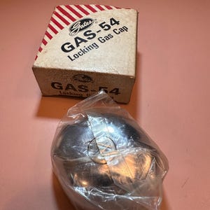 May include: A silver-colored metal locking gas cap enclosed in a clear plastic bag, positioned in front of a white cardboard box. The box features red diagonal stripes and black text that reads Gates GAS-54 Locking Gas Cap on the top and side.
