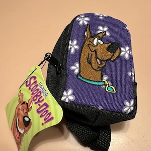 May include: A small, black and purple backpack featuring an embroidered image of Scooby-Doo. The backpack has a purple background with white flower accents. A tag with the Scooby-Doo logo is attached.