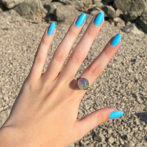 May include: A hand with light blue nail polish is wearing a ring with a green and purple stone. The ring is made of gold wire.