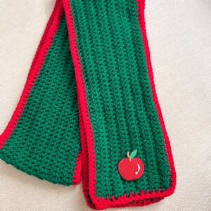 May include: A green crochet scarf with a red border and a red apple applique. The scarf is approximately 60 inches long and 6 inches wide. The apple applique is on one end of the scarf.