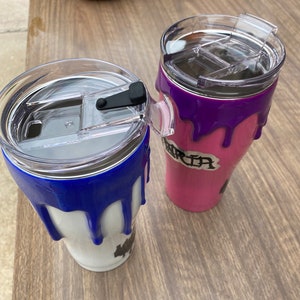 Puede incluir: Dos vasos de acero inoxidable con tapas transparentes. Uno es blanco con pintura azul goteando, el otro es rosa con pintura morada goteando. El vaso rosa tiene el nombre "Maria".