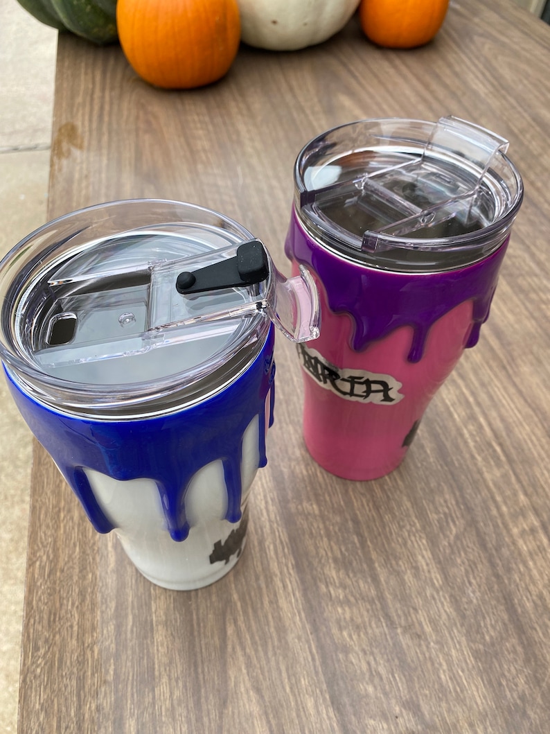 Puede incluir: Dos vasos de acero inoxidable con tapas transparentes. Uno es blanco con pintura azul goteando, el otro es rosa con pintura morada goteando. El vaso rosa tiene el nombre "Maria".