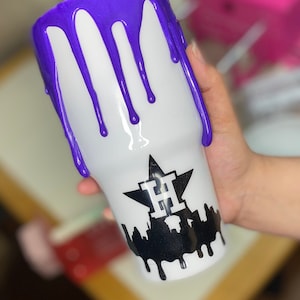 Puede incluir: Vaso blanco con un dise&ntilde;o de goteo morado en la parte superior. El vaso presenta un gr&aacute;fico negro de una estrella con la letra "H" en el interior y una silueta de paisaje urbano con pintura goteando.