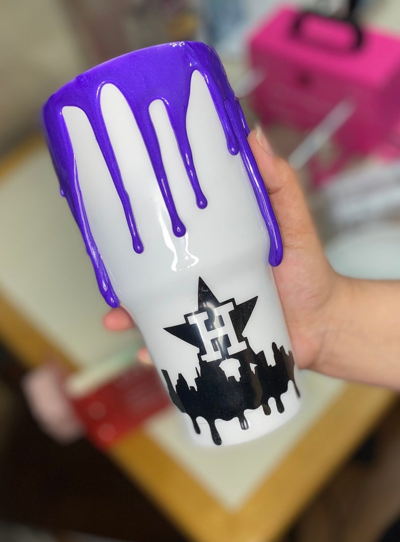Puede incluir: Vaso blanco con un dise&ntilde;o de goteo morado en la parte superior. El vaso presenta un gr&aacute;fico negro de una estrella con la letra "H" en el interior y una silueta de paisaje urbano con pintura goteando.