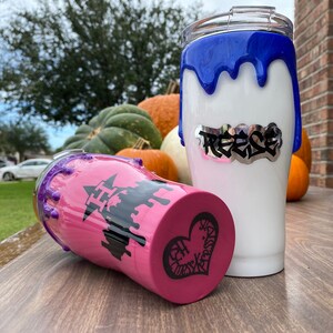 Puede incluir: Dos vasos aislados blancos con dise&ntilde;os de pintura goteada azul y morada. Un vaso tiene el nombre "Reese" en letras plateadas. El otro vaso tiene una base rosa con un gr&aacute;fico de castillo negro y un dise&ntilde;o de coraz&oacute;n.