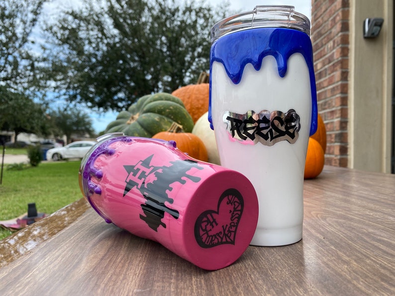 Puede incluir: Dos vasos aislados blancos con dise&ntilde;os de pintura goteada azul y morada. Un vaso tiene el nombre "Reese" en letras plateadas. El otro vaso tiene una base rosa con un gr&aacute;fico de castillo negro y un dise&ntilde;o de coraz&oacute;n.