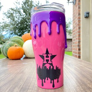 Puede incluir: Un vaso rosa con un dise&ntilde;o de goteo p&uacute;rpura. El vaso presenta un gr&aacute;fico negro de una estrella con la letra "H" y un horizonte de la ciudad. El vaso tiene una tapa transparente.