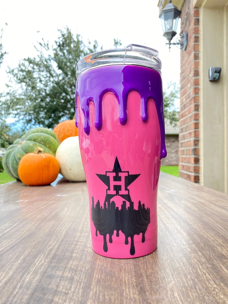Puede incluir: Un vaso rosa con un dise&ntilde;o de goteo p&uacute;rpura. El vaso presenta un gr&aacute;fico negro de una estrella con la letra "H" y un horizonte de la ciudad. El vaso tiene una tapa transparente.